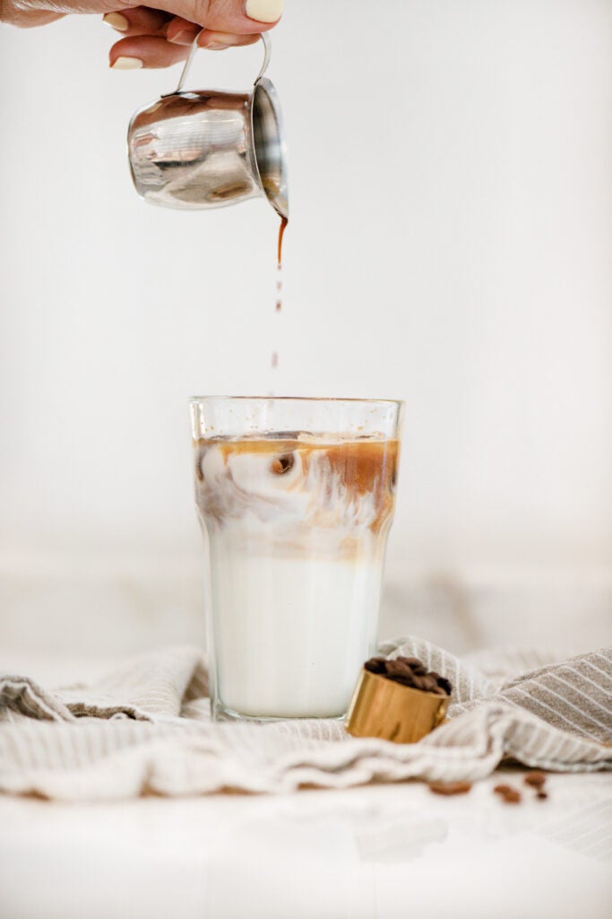 someone pouring espresso into cup of milk on table with decorative towel and coffee beans